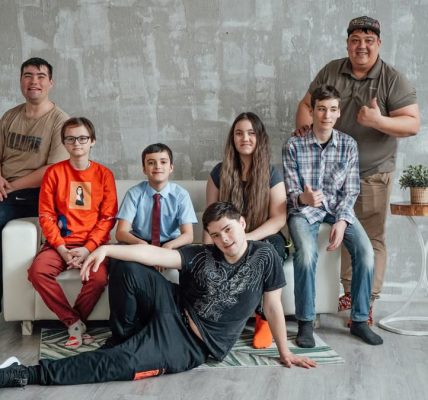 Group of seven people posing casually in a stylish indoor setting with a concrete wall background.
