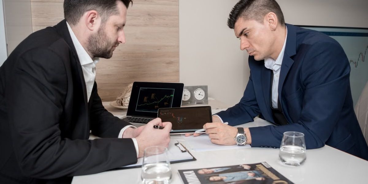 Two businessmen in suits discussing cryptocurrency trends using digital devices in an office setting.