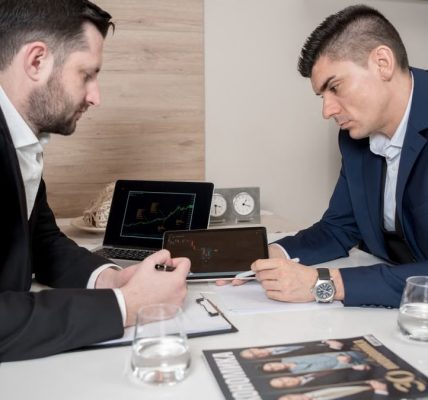 Two businessmen in suits discussing cryptocurrency trends using digital devices in an office setting.