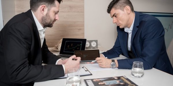 Two businessmen in suits discussing cryptocurrency trends using digital devices in an office setting.