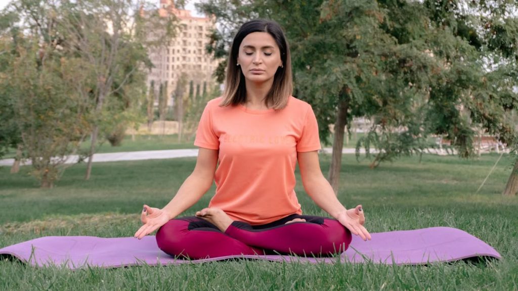 Woman meditating in lotus position on a yoga mat in a sunny park.